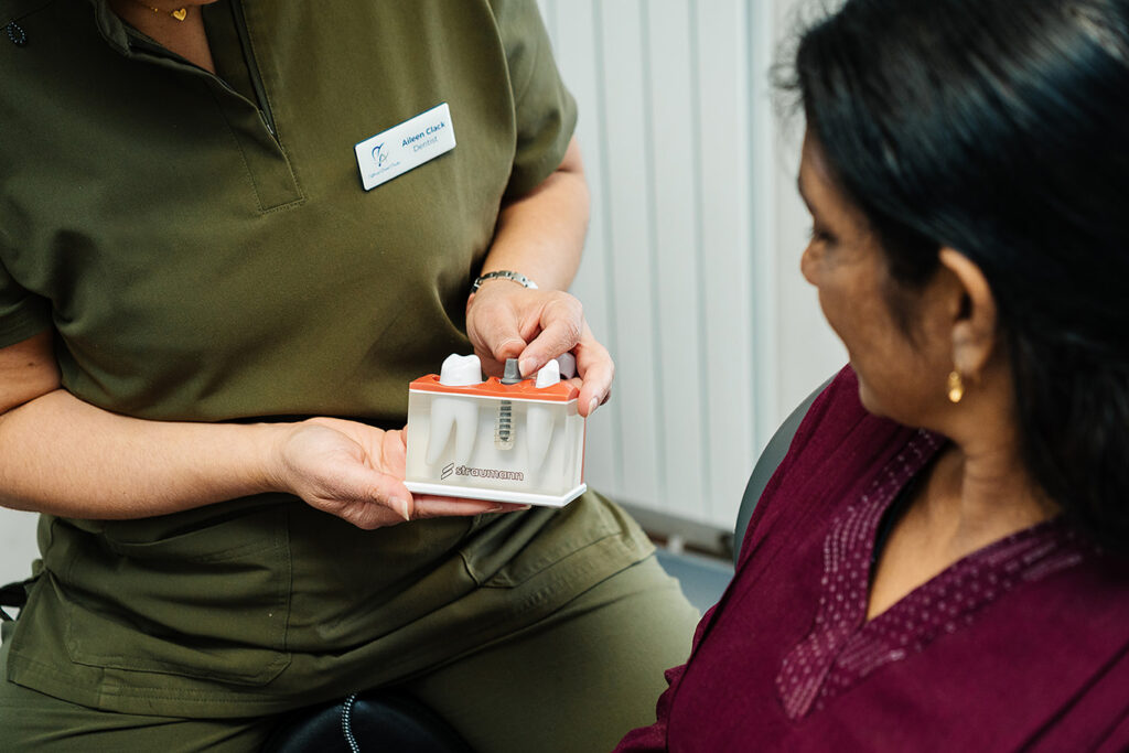 A Toftwood Dental Practice's dentist in olive green scrubs, holds a dental model showing a dental implant. The model includes artificial teeth and a visible metal implant screw.