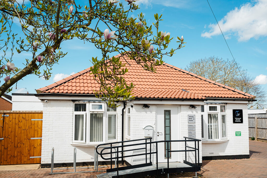Exterior view of Toftwood Dental Practice, a white single-storey building with a red tiled roof, accessible ramp, and spring blossoms in the foreground.