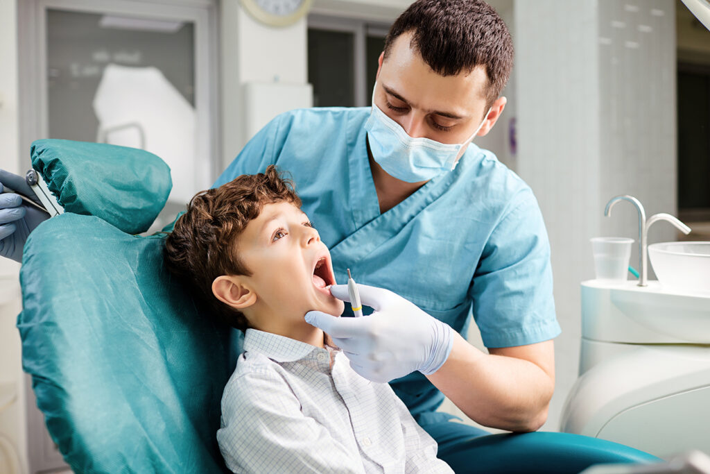 Male dentist in a blue uniform and face mask examining a young boy's teeth in a dental chair, creating a calm and reassuring environment during a routine check-up.