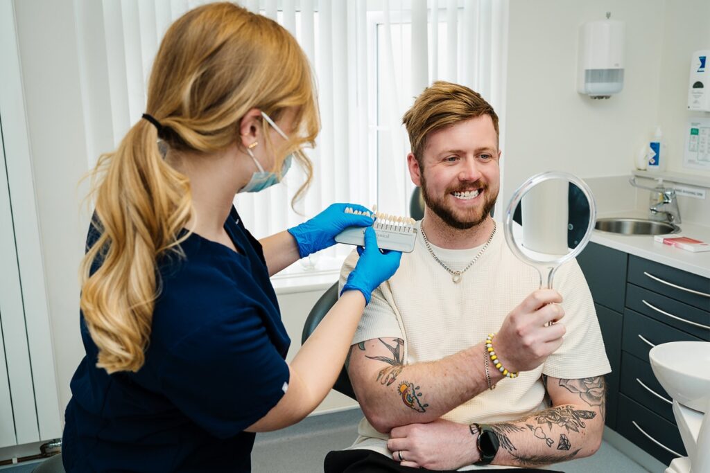 A female dentist holds up a shade guide to compare tooth colors while a smiling male patient looks into a handheld mirror.